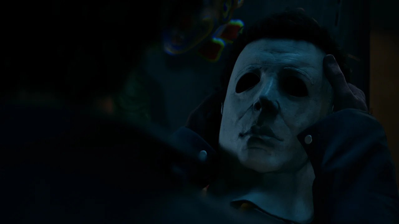 Halloween: The Game screenshot: Michael Myers in his mask standing in a dark Haddonfield kitchen holding a knife, lit by a single light source